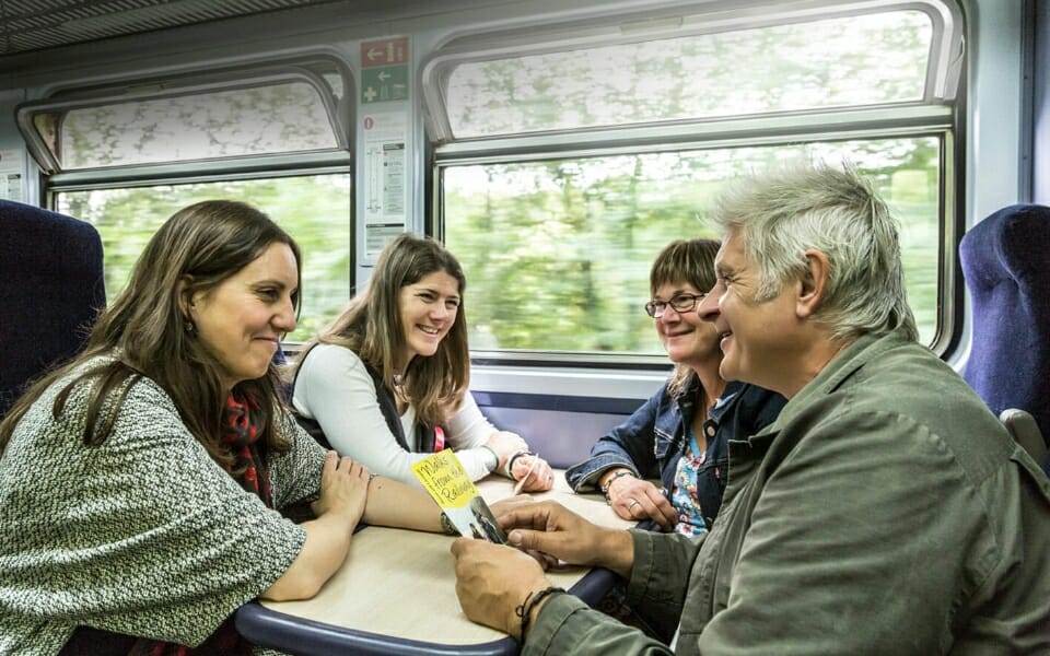 four passengers smiling sat at table seats on a train with one holding a destination guide