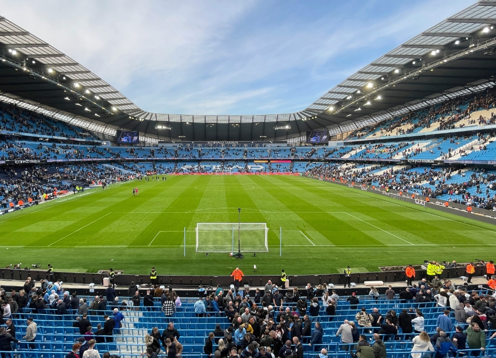 The Manchester City Etihad Stadium with tiered blue seats with fans standing and a grass football pitch in the centre