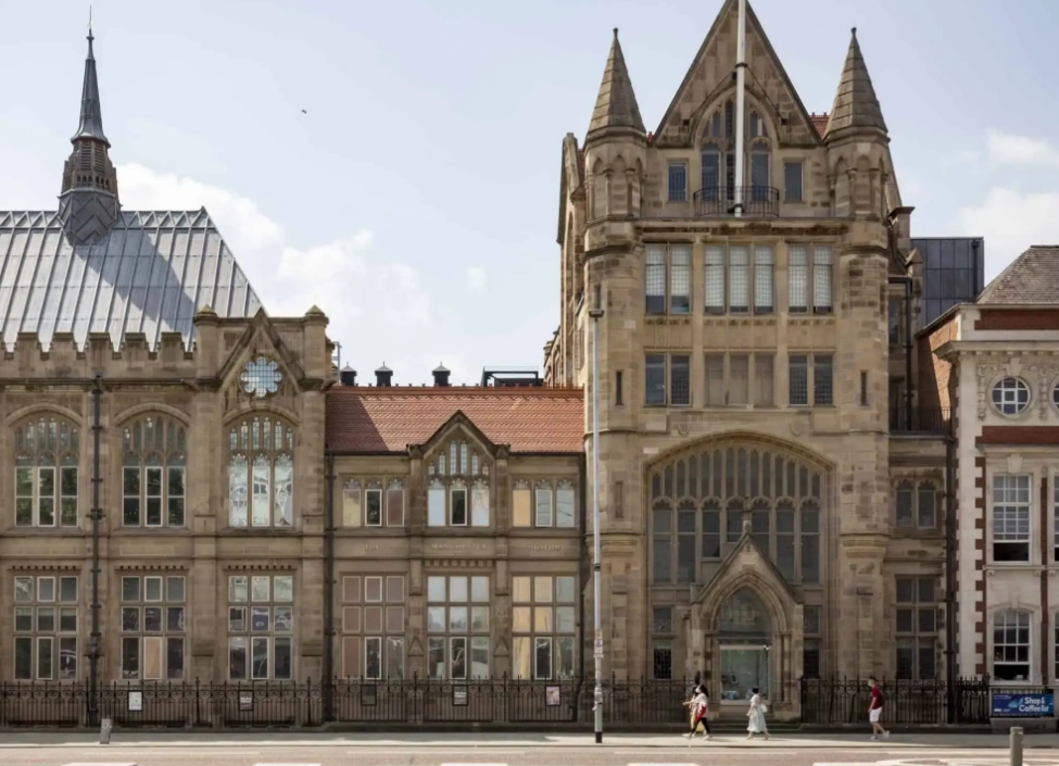 The Manchester Museum, an old windowed neo-gothic building with people walking past and a road in front