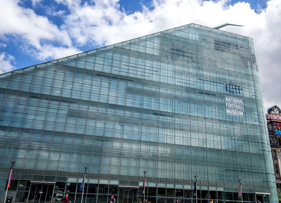 The National Football Museum, a tall glass windowed building of a geometric shape with a pavement in front and people walking past